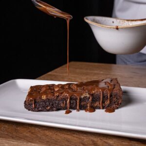 Brownie tradicional de chocolate servido en porción rectangular, cubierto con salsa de chocolate caliente sobre plato blanco.