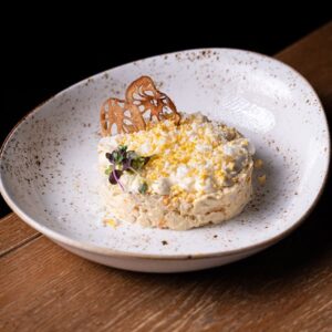 Ensaladilla rusa cremosa presentada en forma circular, decorada con huevo rallado y brotes, servida en plato hondo blanco.