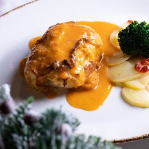 Merluza rellena de centollo cubierta con salsa de carabineros, servida en plato con guarnición de patata y brócoli.