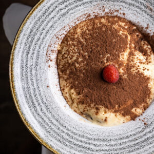 Tiramisú casero con cacao espolvoreado y una frambuesa en el centro, servido en plato blanco visto desde arriba.