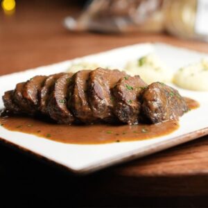 Carrilleras guisadas al vino tinto cortadas en medallones, servidas con su salsa en plato rectangular.